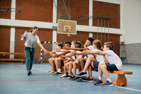 Happy sports teacher greeting with group of elementary students during PE class at school gym.