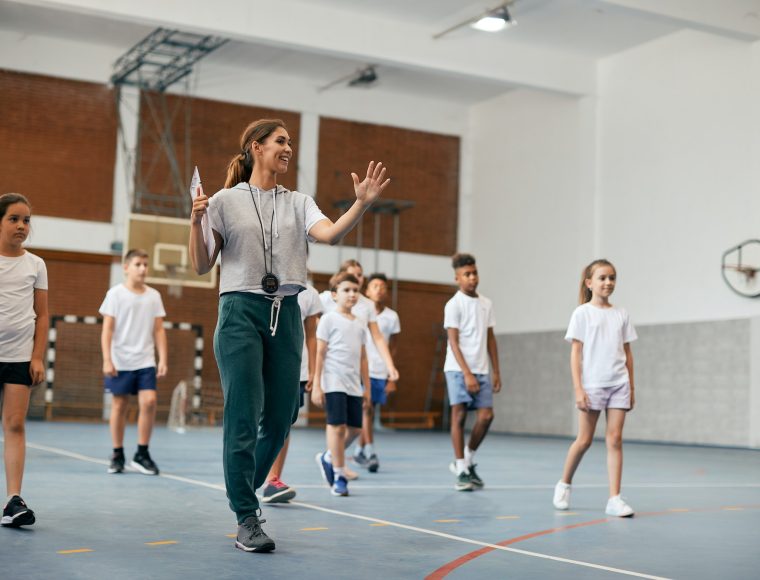Young female coach having PE class with group of elementary students at school gym.
